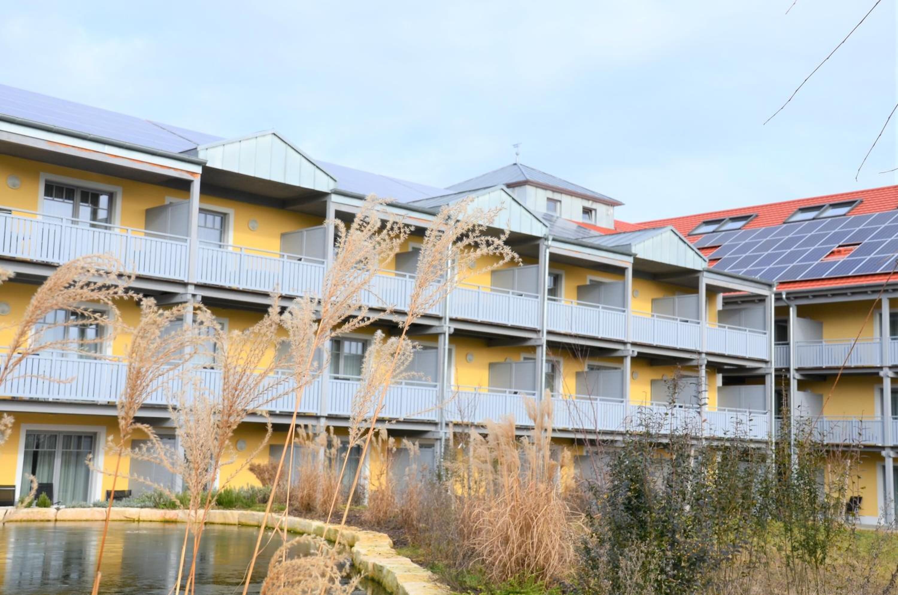 Un edificio de varios pisos con balcones, paneles solares en el techo y un pequeño estanque al frente.