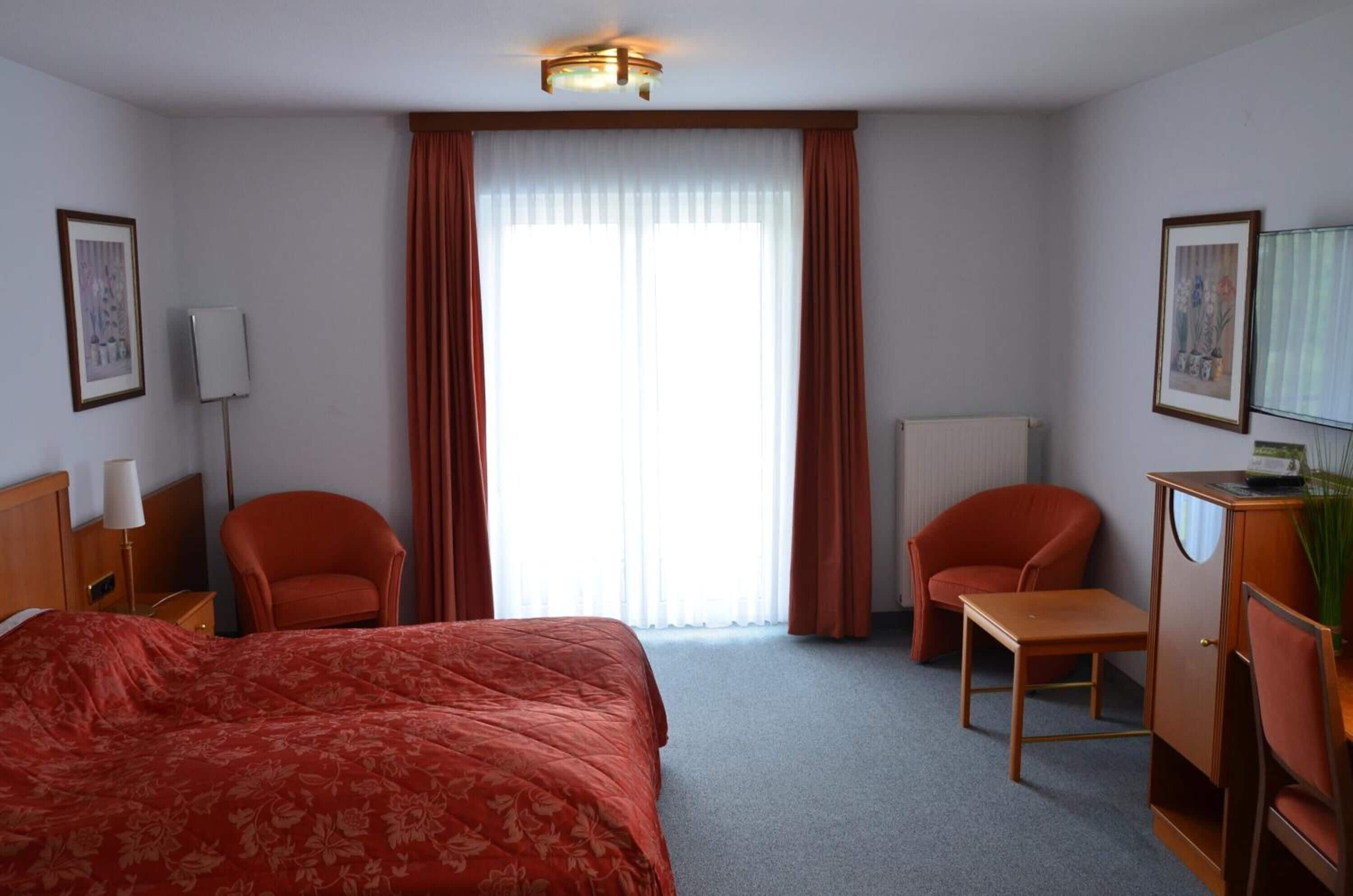 Habitación de hotel con cama, dos sillas, escritorio, radiador y ventana con cortinas.