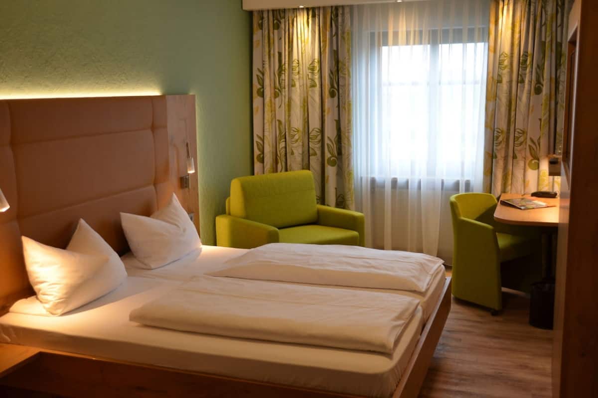 A hotel room with a large bed, two green chairs, a small table, and a window with curtains.