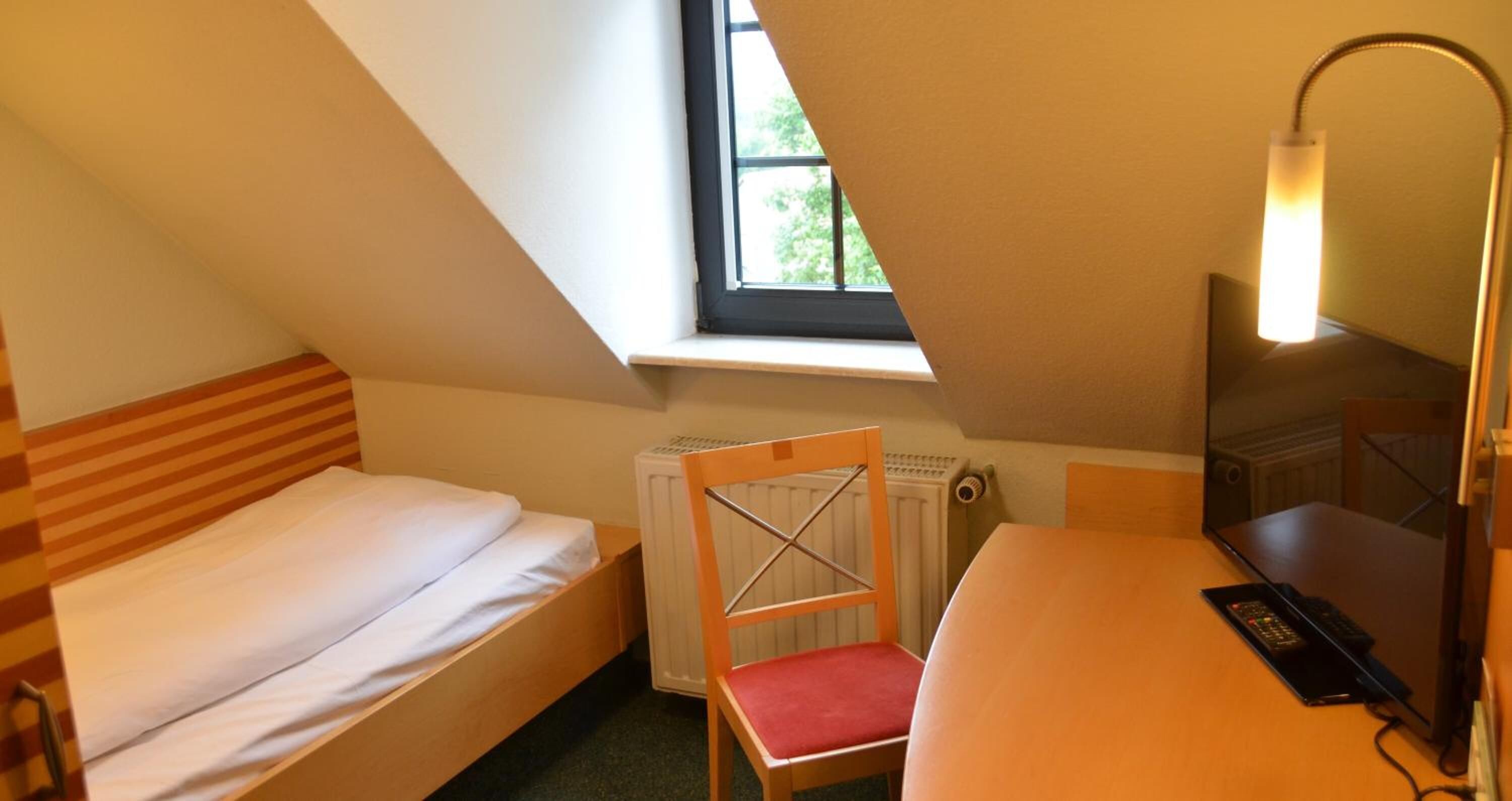 Una habitación pequeña y triangular con una cama individual, una silla, un escritorio y una lámpara.