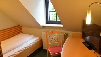 Una habitación pequeña y triangular con una cama individual, una silla, un escritorio y una lámpara.