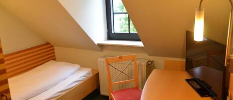 Ein kleiner, dreieckiger Raum mit einem Einzelbett, einem Sessel, einem Schreibtisch und einer Lampe.
