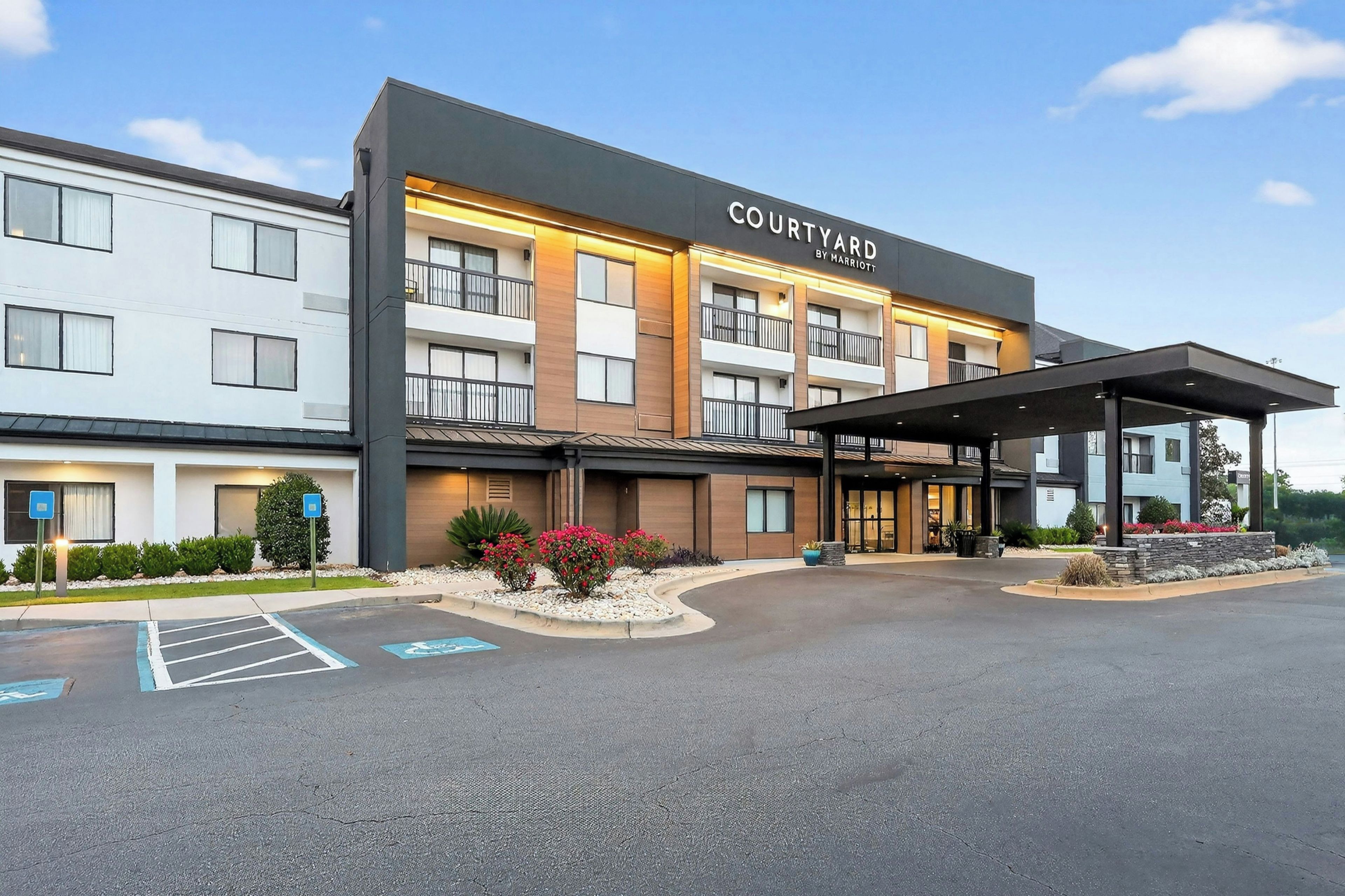 Ein Courtyard by Marriott Hotel mit überdachter Zufahrt, Parkplatz und einer begrünten Anlage mit Sträuchern und Blumen.