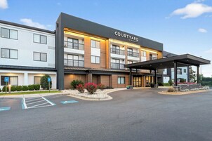 Un hotel Courtyard by Marriott con entrada cubierta, estacionamiento y un área ajardinada con arbustos y flores.
