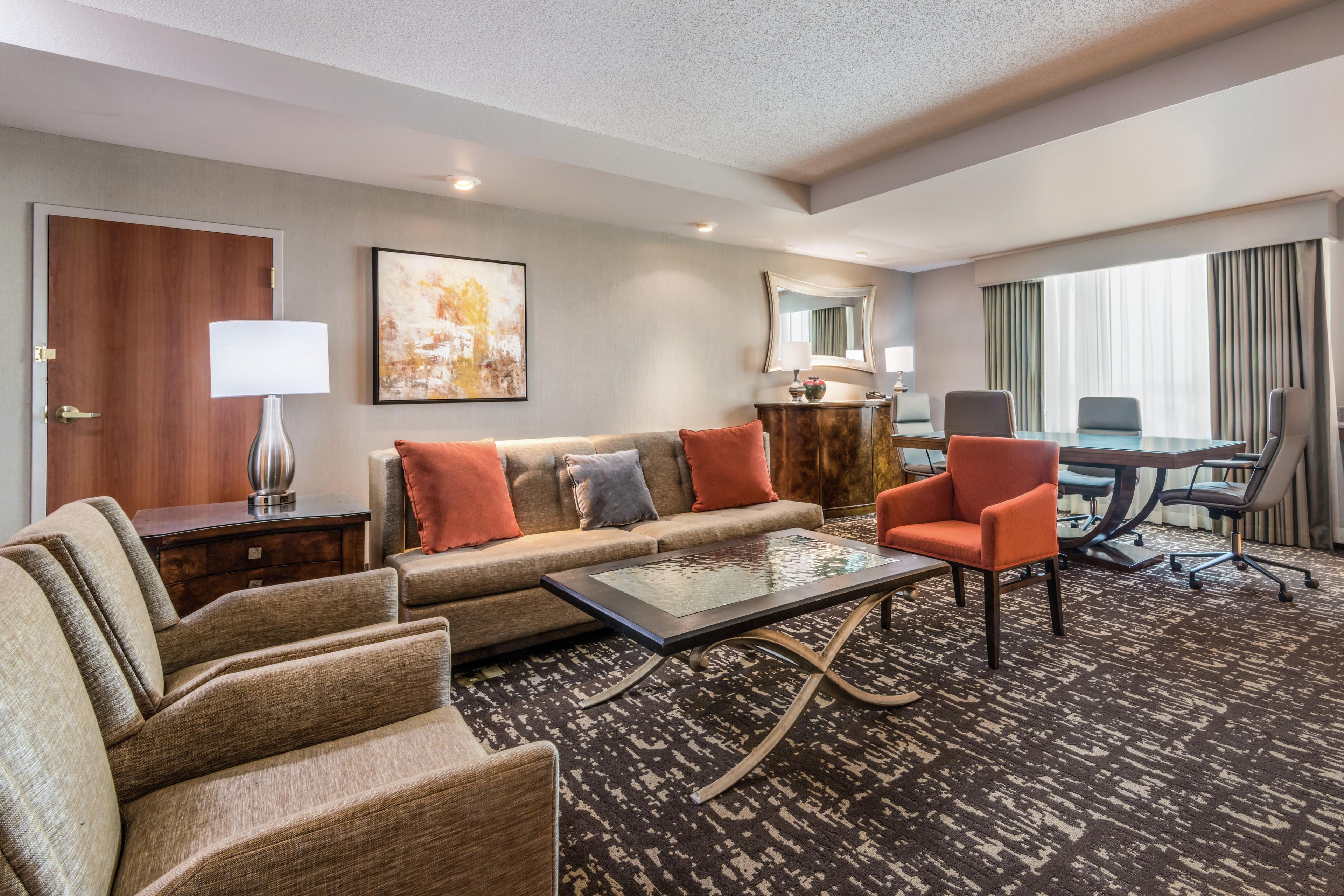 A hotel room with a sofa, armchairs, a coffee table, a desk with a chair, a lamp, a painting, and a large window with curtains.