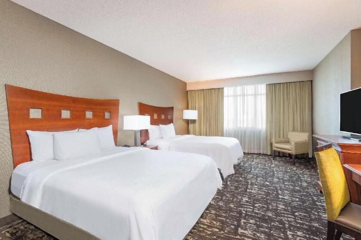A hotel room with two beds, a desk with a chair, a television, and a large window with curtains.