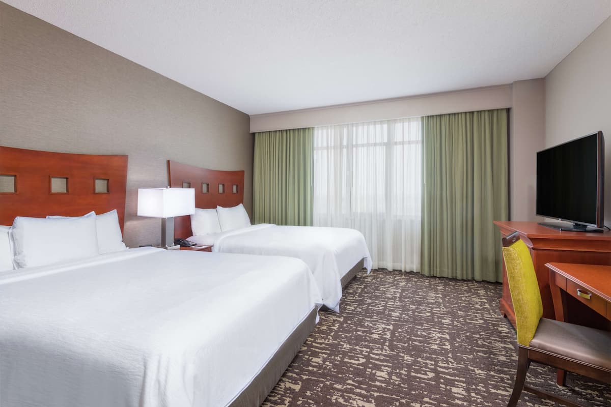 Habitación de hotel con dos camas, un escritorio con televisor, una silla y una ventana con cortinas.