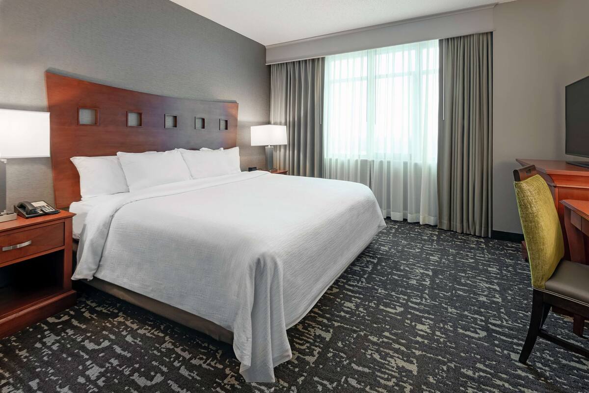 Habitación de hotel con una cama grande, un escritorio con silla, televisor, mesita de noche y ventana con cortinas.