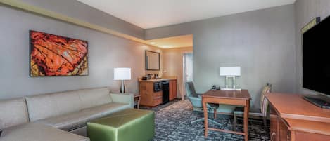 Habitación de hotel con sofá, un otoman verde, una mesa de comedor con sillas, televisión y minibar.