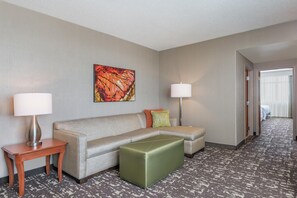 A hotel room with a sofa, a small table, a green ottoman, a painting on the wall, and a lamp. - Embassy Suites by Hilton St. Louis St. Charles (St. Charles)