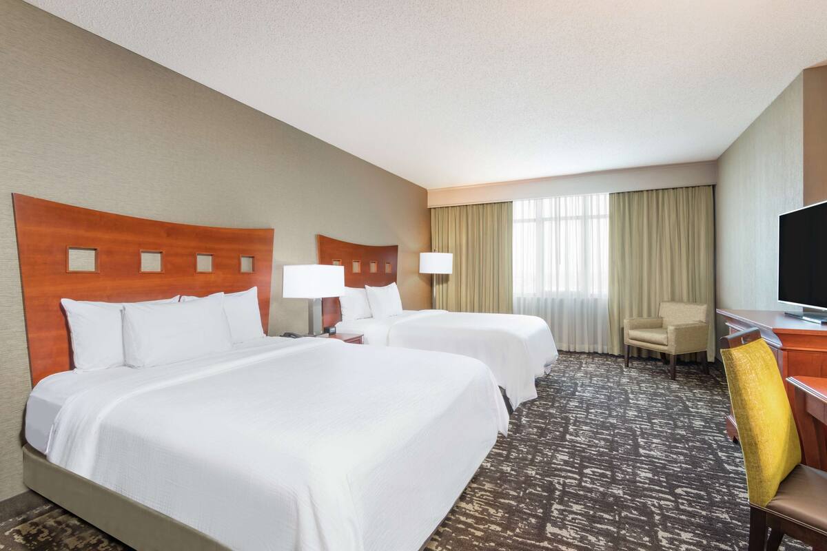 Habitación de hotel con dos camas, un escritorio, una silla, un televisor y un ventanal grande con cortinas.