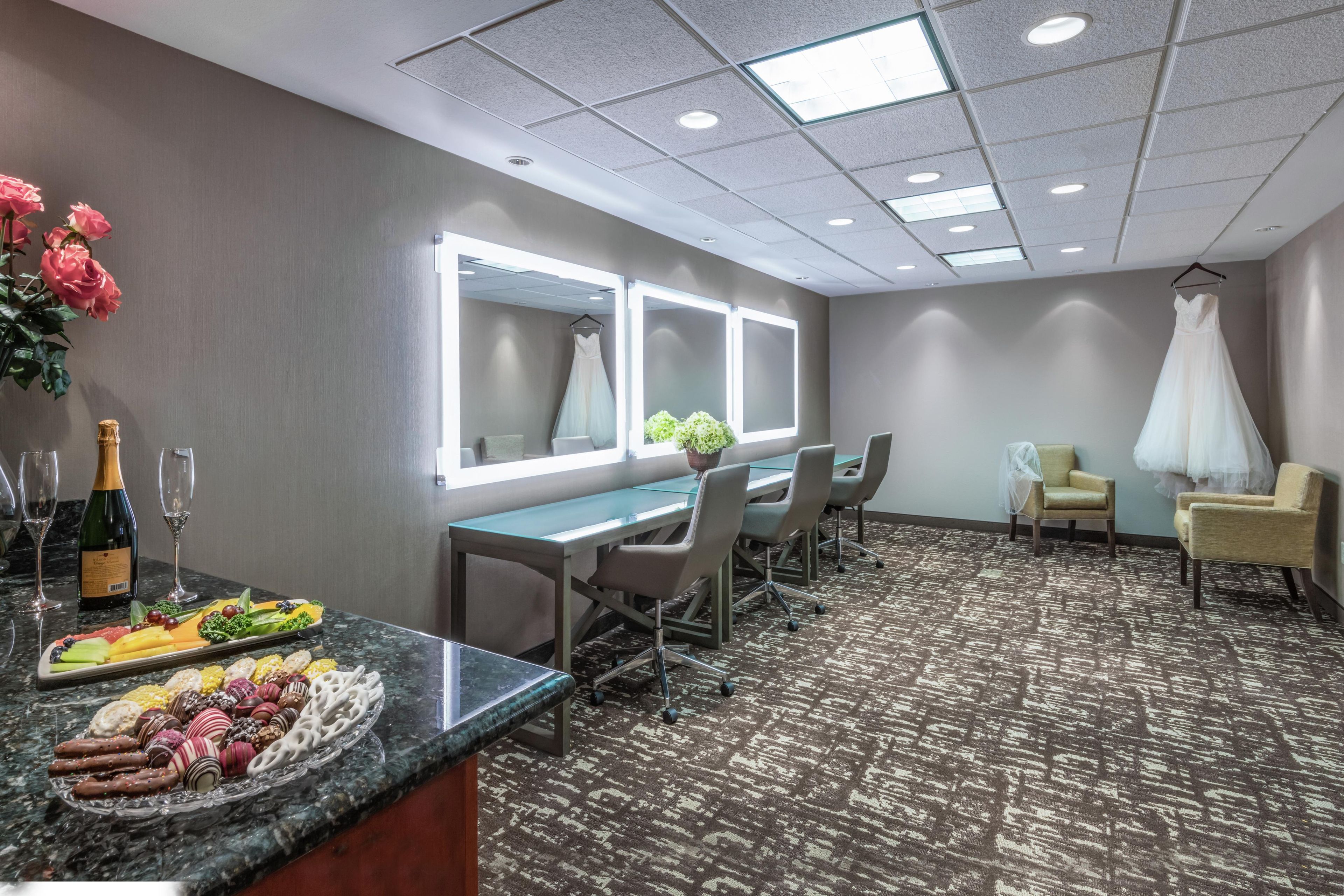 A hotel room with a dining area, a mirror, a dress on a chair, and a carpeted floor.