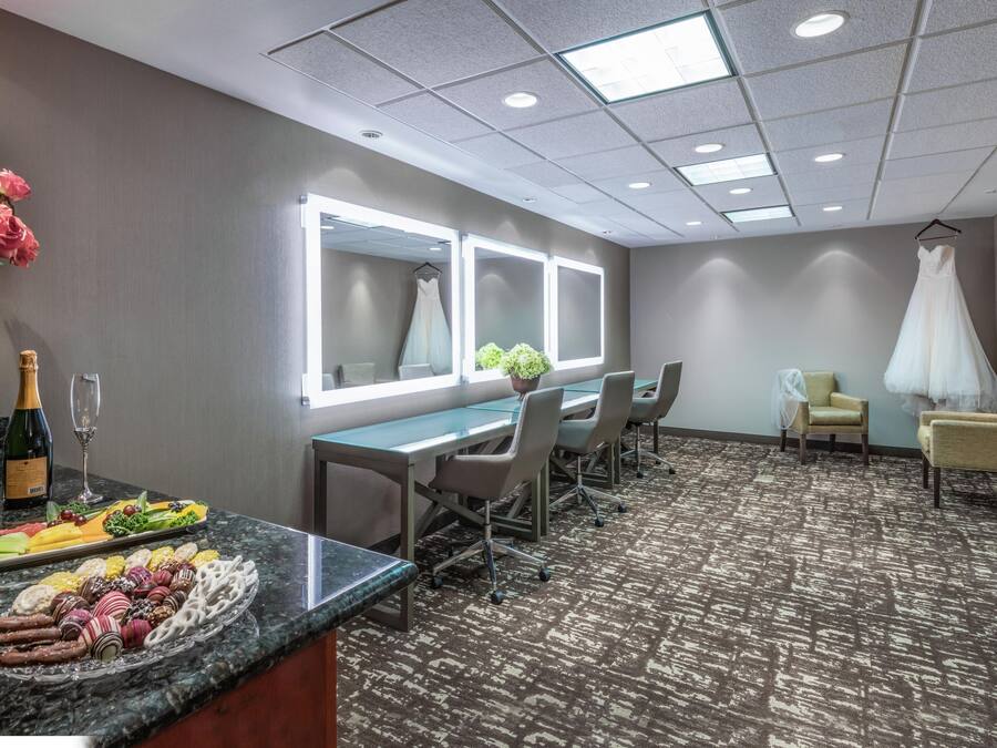 A hotel room with a dining area, a mirror, a dress on a chair, and a carpeted floor.