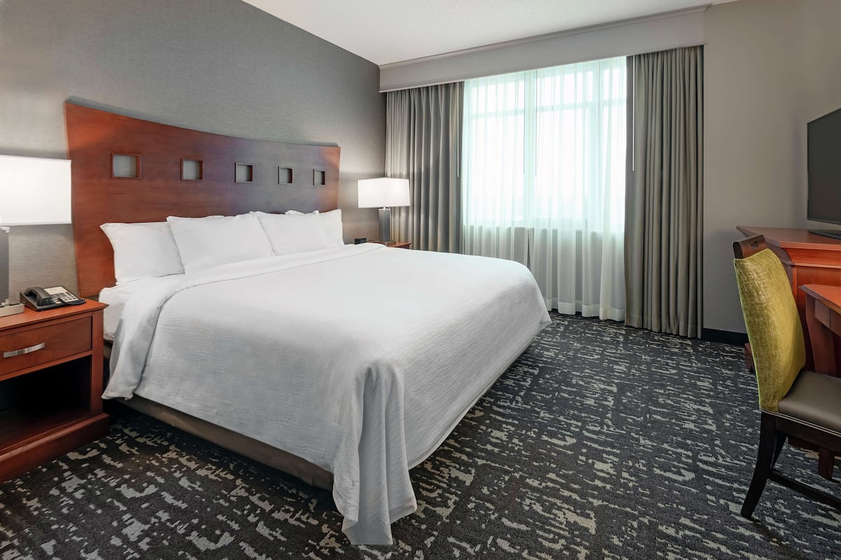 Habitación de hotel con una cama grande, un escritorio con una silla, televisión y ventana con cortinas.