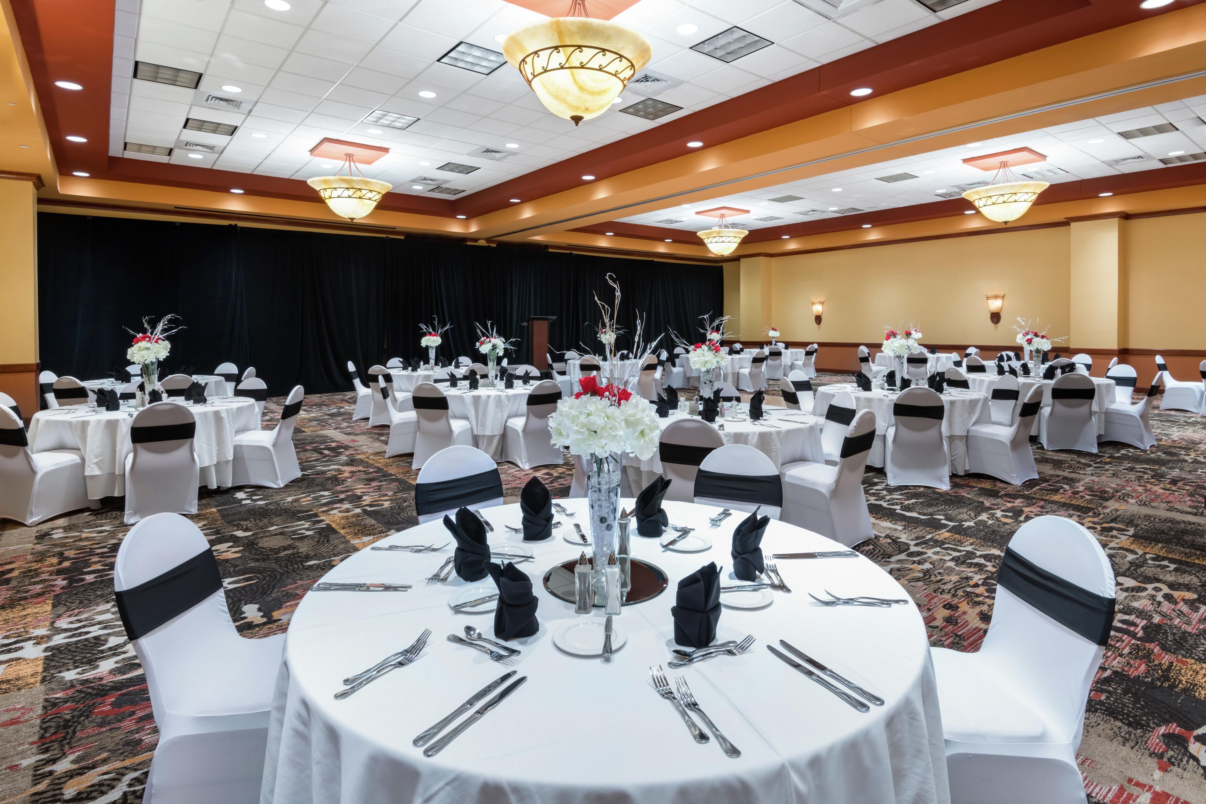 Un salón de banquetes con mesas redondas dispuestas para un evento formal, con manteles blancos, fundas negras para las sillas y centros de mesa florales.