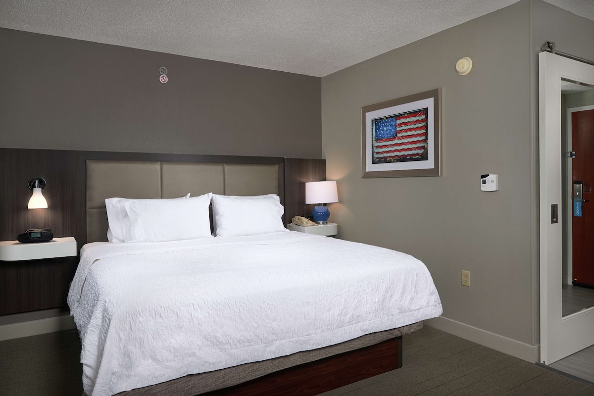 Habitación de hotel con una cama grande, una mesita de noche con lámpara, una bandera estadounidense enmarcada y una puerta a la derecha.