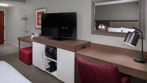 A hotel room with a bed, a desk with a television, a red chair, a mirror, and a lamp. - Hampton Inn & Suites Oxford Anniston (Oxford)