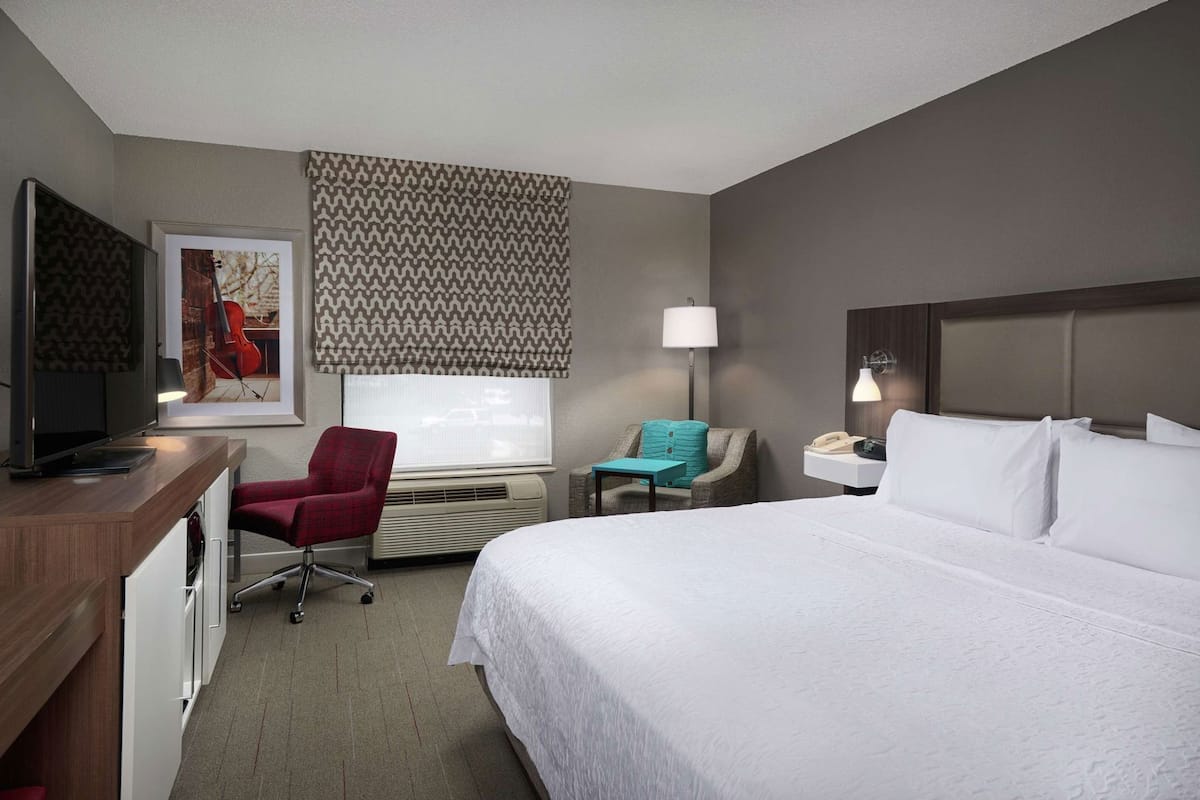 Habitación de hotel con cama, escritorio con televisor, una silla, mesita de noche con lámpara y ventana con persiana decorativa.