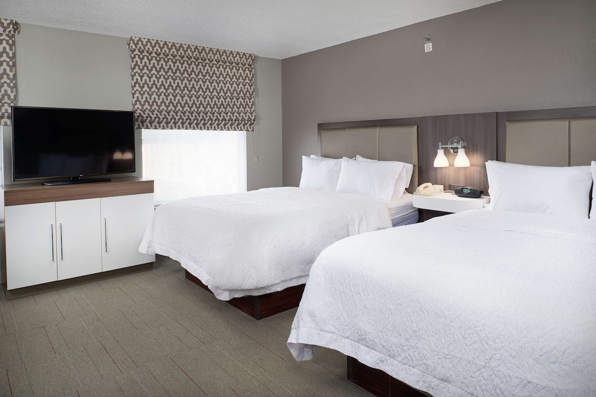 Habitación de hotel con dos camas, un televisor, una mesita de noche con lámpara y una ventana con cortinas estampadas.
