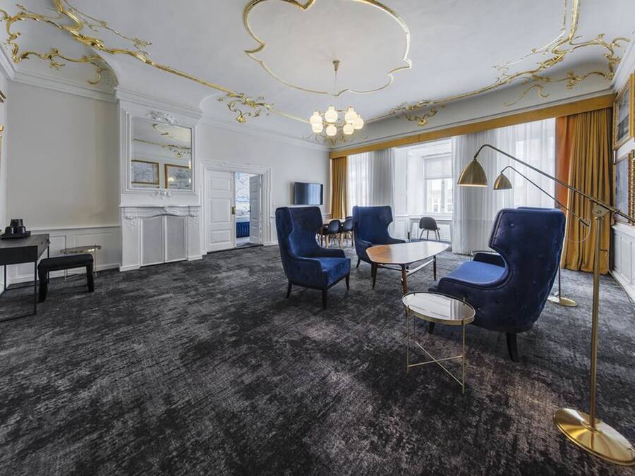 A room with a dark carpet, blue armchairs, a round table, and a gold floor lamp.