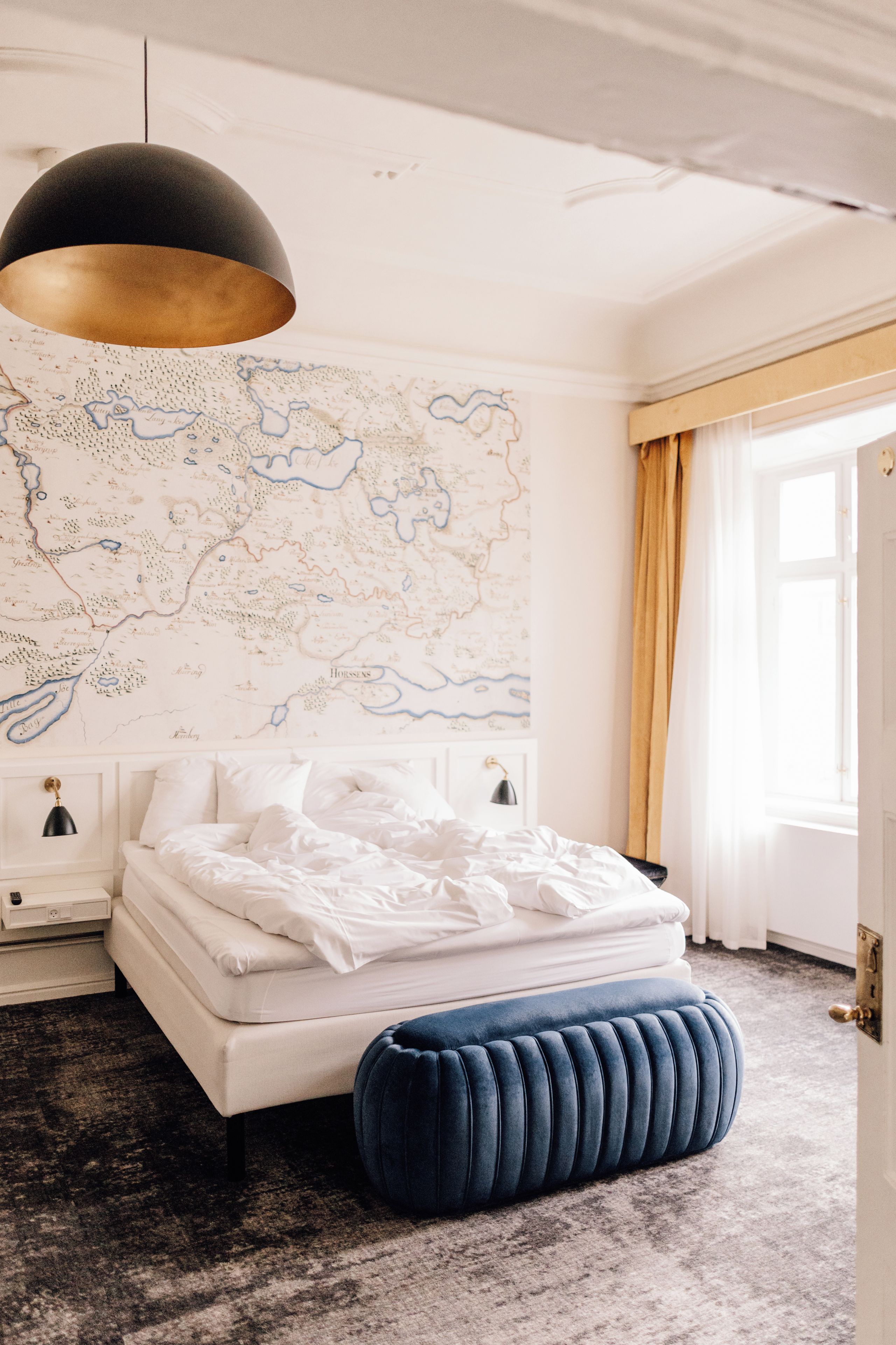 Un dormitorio con cama, un mapa en la pared, una mesita de noche, una lámpara y una ventana con cortinas.