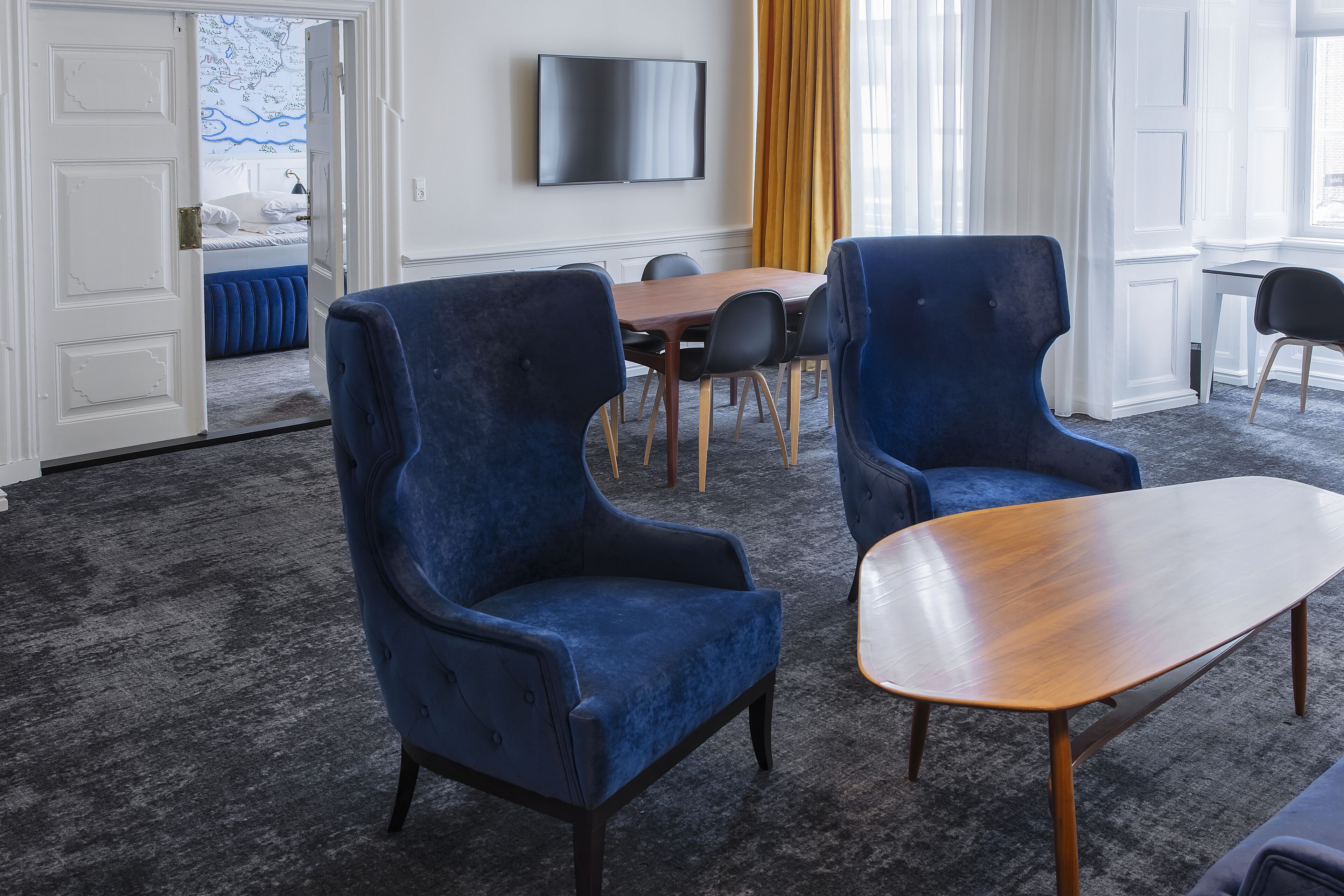 A modern hotel room with a large bed, a dining table, two blue armchairs, a wooden coffee table, and a flat-screen TV on the wall.