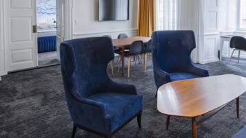 A modern hotel room with a large bed, a dining table, two blue armchairs, a wooden coffee table, and a flat-screen TV on the wall.