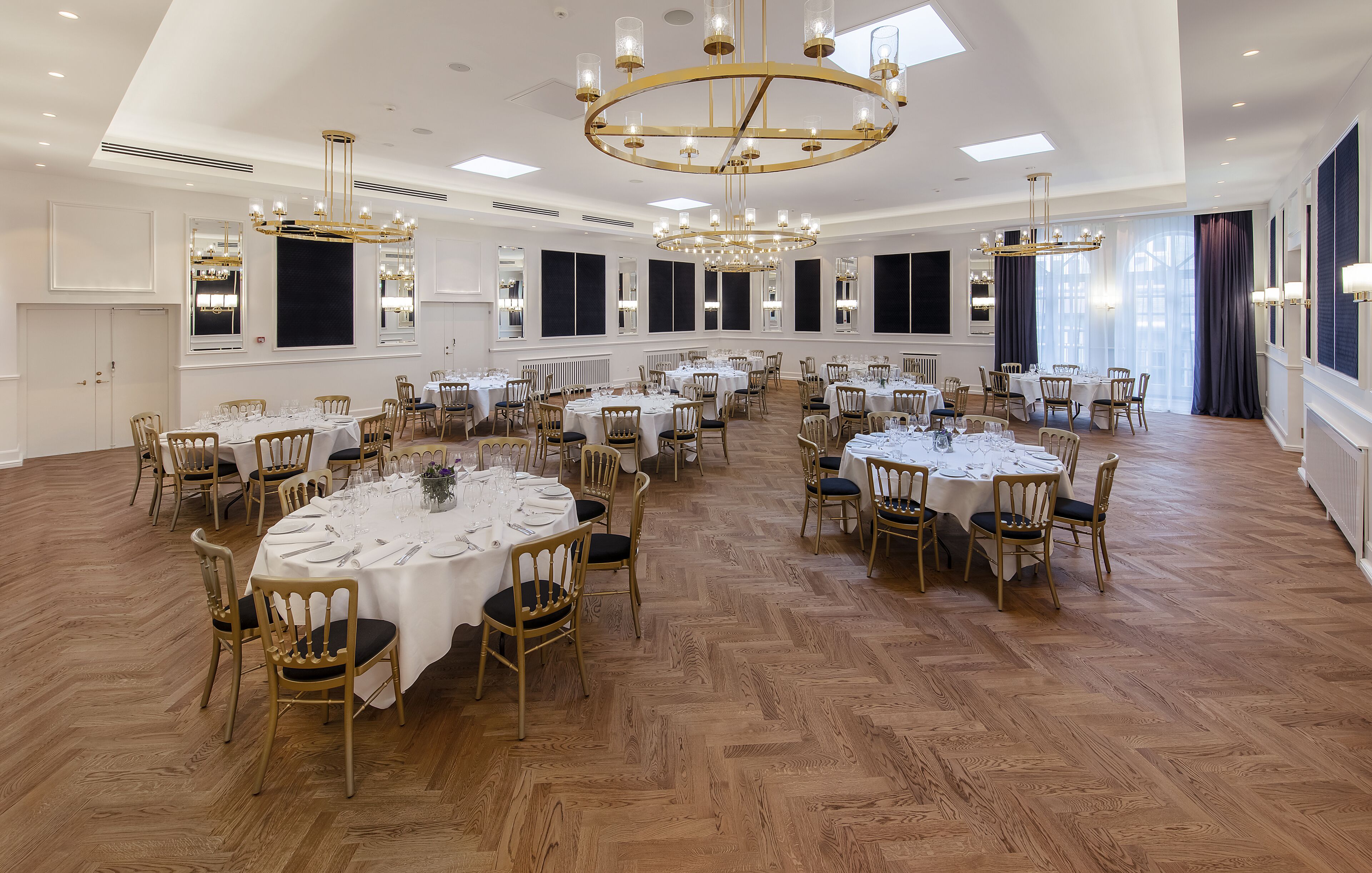 Un amplio salón de banquetes con varias mesas redondas, sillas doradas y elegantes lámparas de araña.