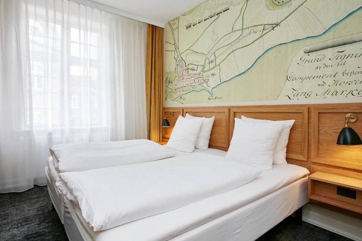 Une chambre d’hôtel avec deux lits, une tête de lit en bois, une carte accrochée au mur et une fenêtre ornée de rideaux légers.