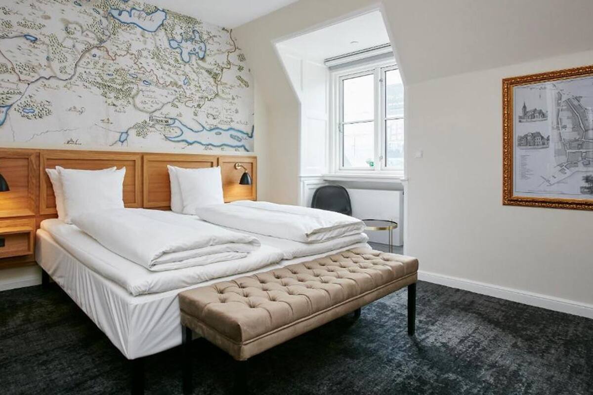 Une chambre d’hôtel avec un grand lit, un banc, une carte accrochée au mur et une fenêtre.