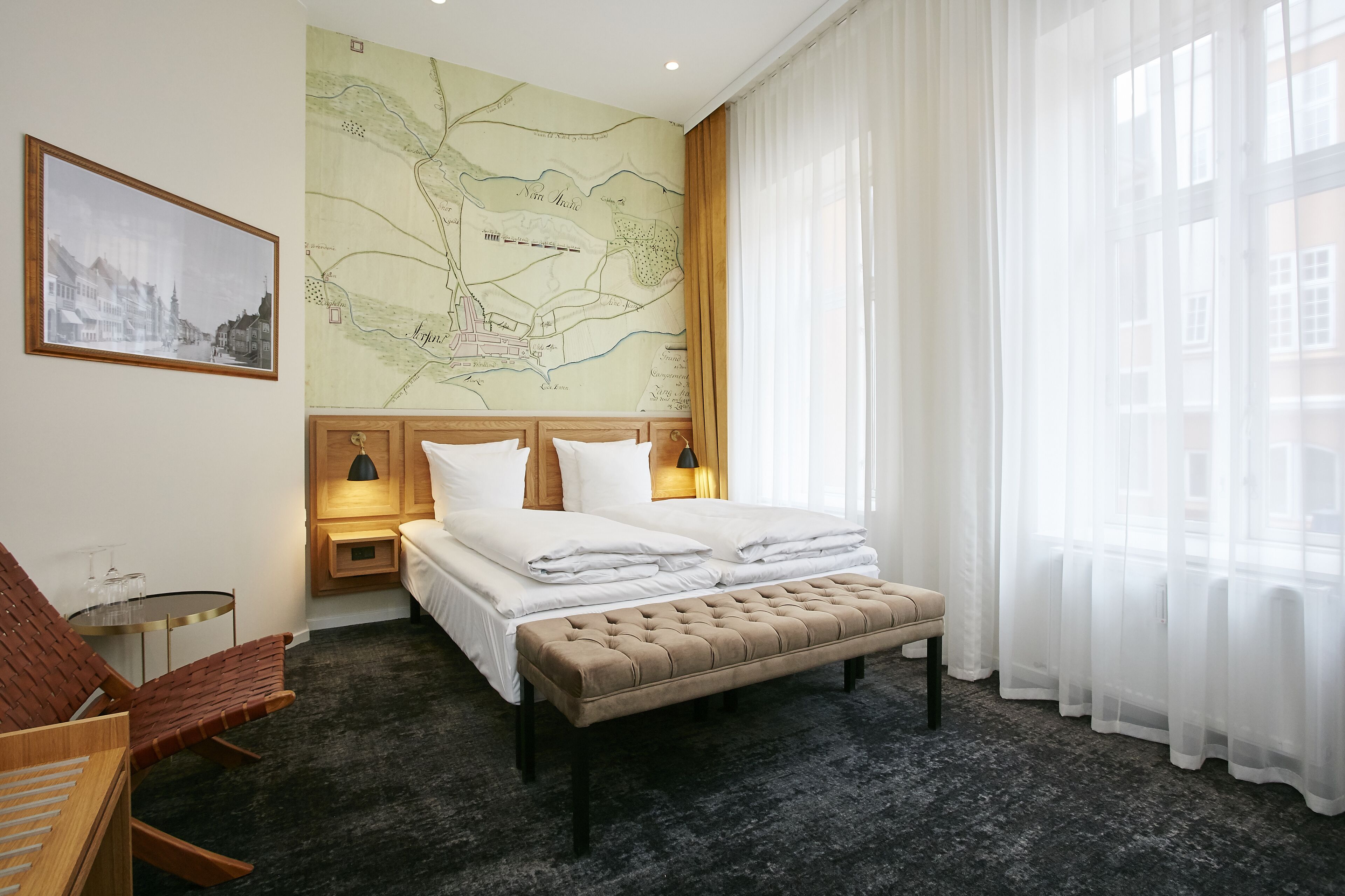 A neatly arranged hotel room with a bed, a map on the wall, a chair, and a bench.