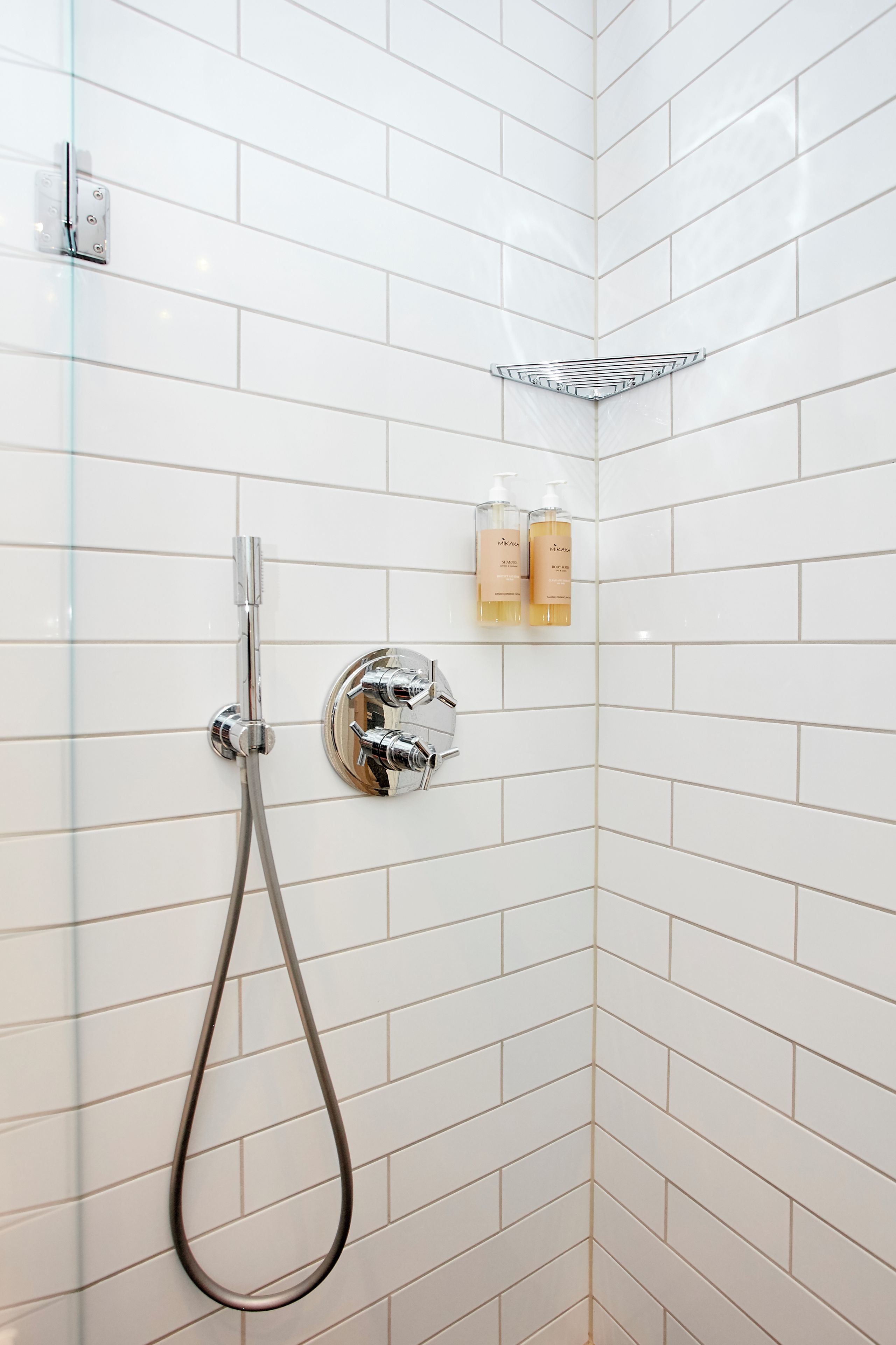 En bruser med kromarmatur og glasdør, hvid klinkevæg og to shampoo-flasker på en hylde.