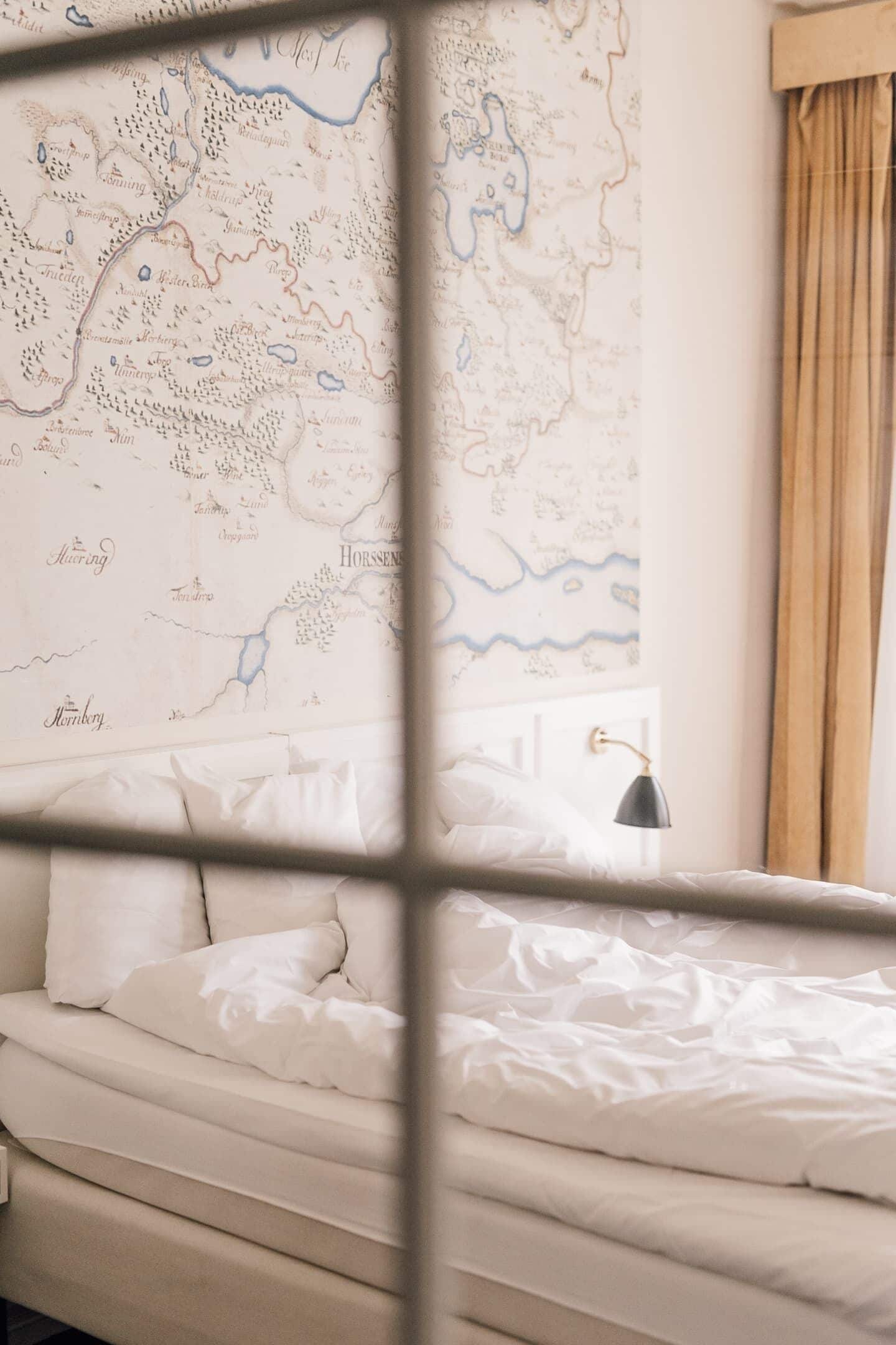 A bed with white bedding, a map on the wall, and a window with curtains.
