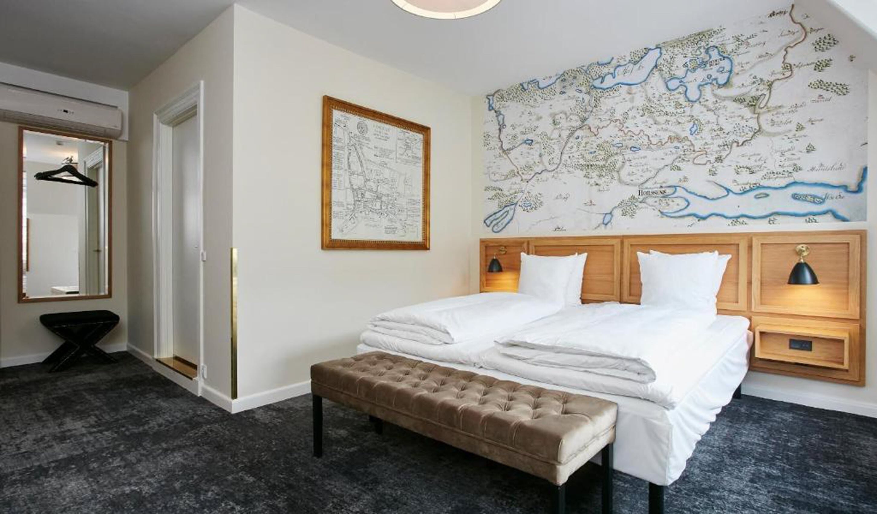 Habitación de hotel con un mapa grande en la pared, una cama con sábanas blancas, un banco y una mesita de noche con lámparas.