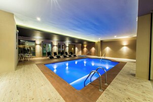 An indoor swimming pool with a blue water feature, surrounded by lounge chairs and a seating area with tables.