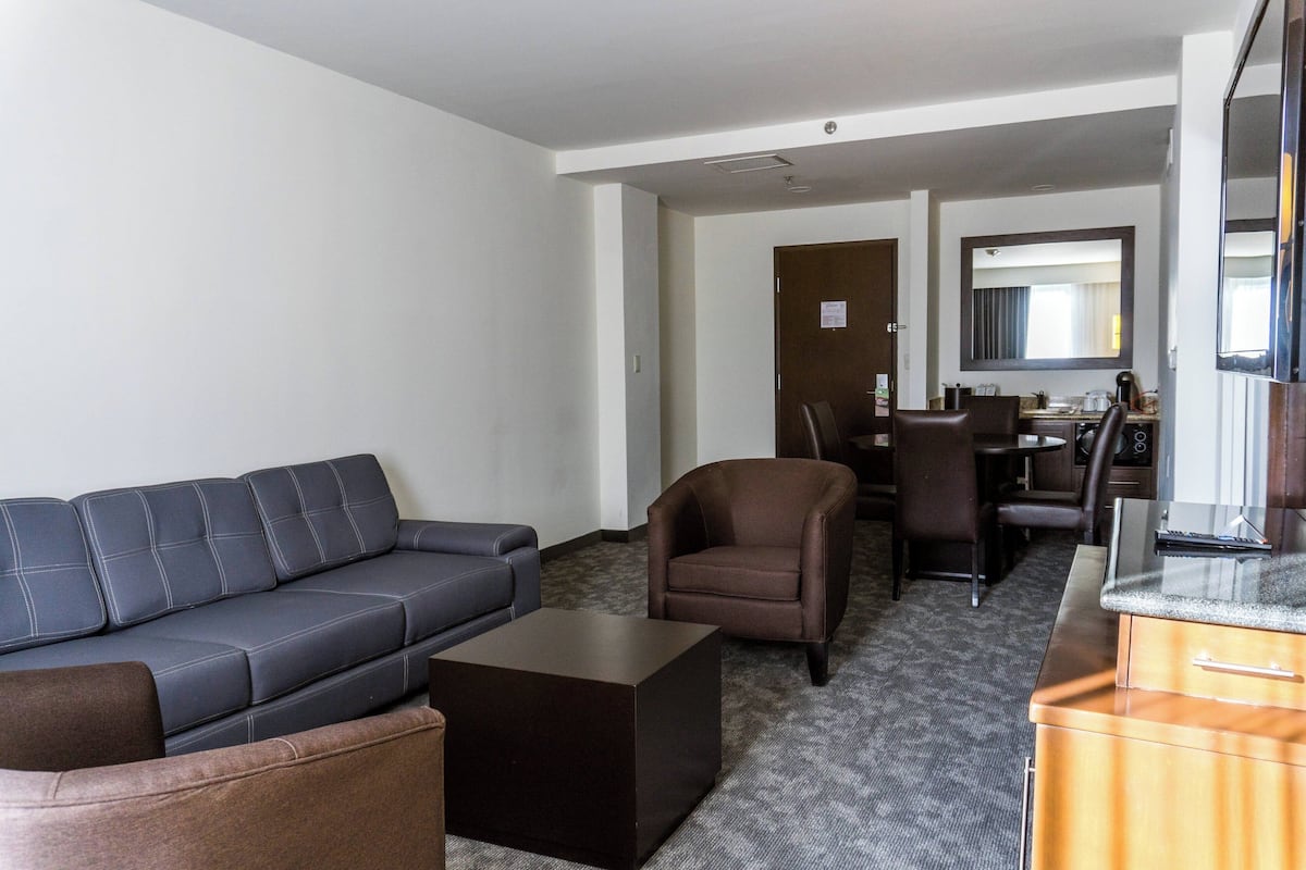 Une chambre d’hôtel avec un canapé gris foncé, un fauteuil marron, une petite table en bois et un coin cuisine équipé d’un miroir et d’un plan de travail.