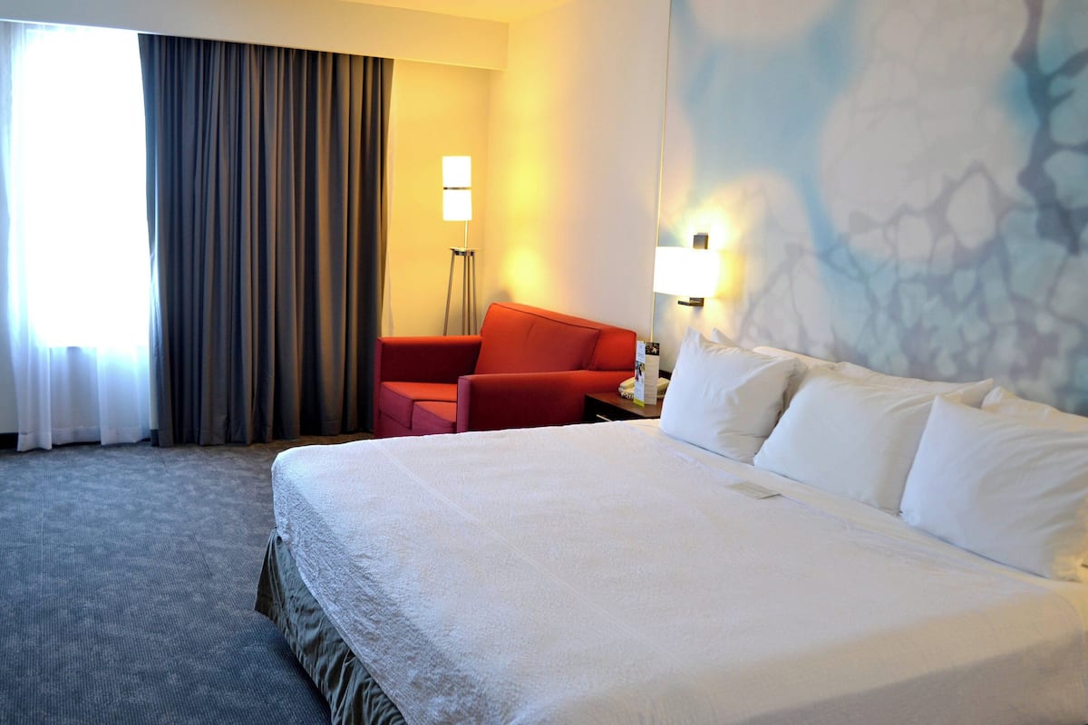 Une chambre d’hôtel avec un grand lit, un fauteuil rouge, un lampadaire et une vue sur l’extérieur par une fenêtre avec des rideaux.