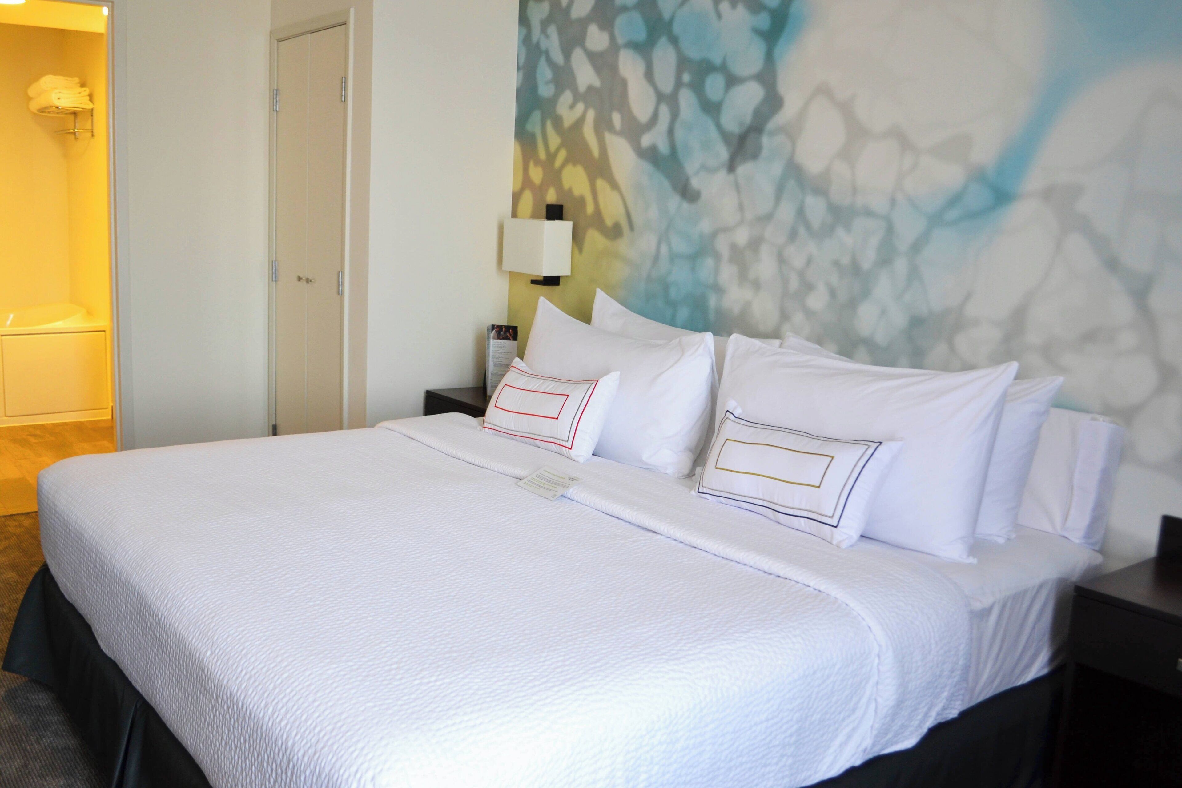 Habitación de hotel con una cama grande, una mesita de noche, una lámpara de pared y un baño visible a la izquierda.