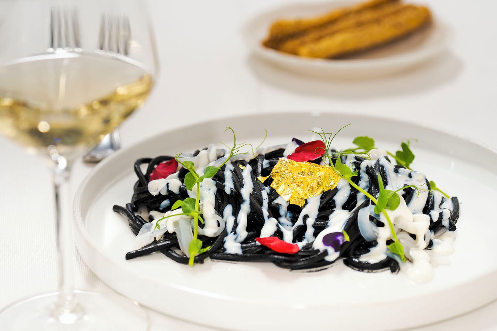 Eine Schüssel schwarze Pasta mit weißer Sauce, garniert mit Grünzeug und Blattgold, dazu ein Glas Weißwein.