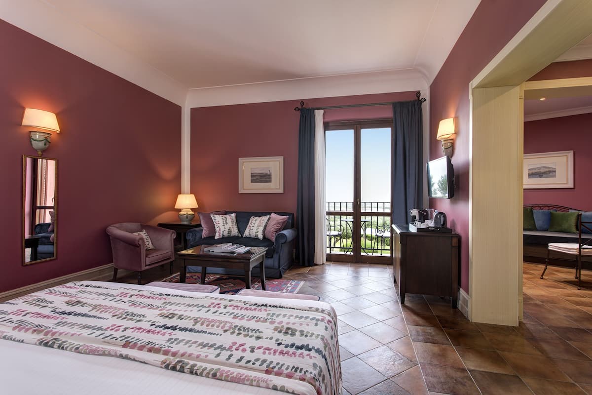 Habitación de hotel con una cama grande, un sofá, una mesita, un televisor y un balcón con vistas.