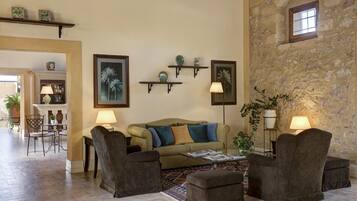 A cozy living room with a sofa, armchairs, and a coffee table. There are wall-mounted shelves with decorative items and a stone wall with a window.