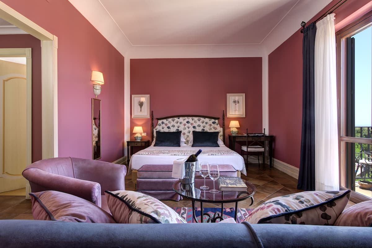 Un dormitorio con una cama, una silla, una mesa con una botella y vasos, y una ventana con cortinas.