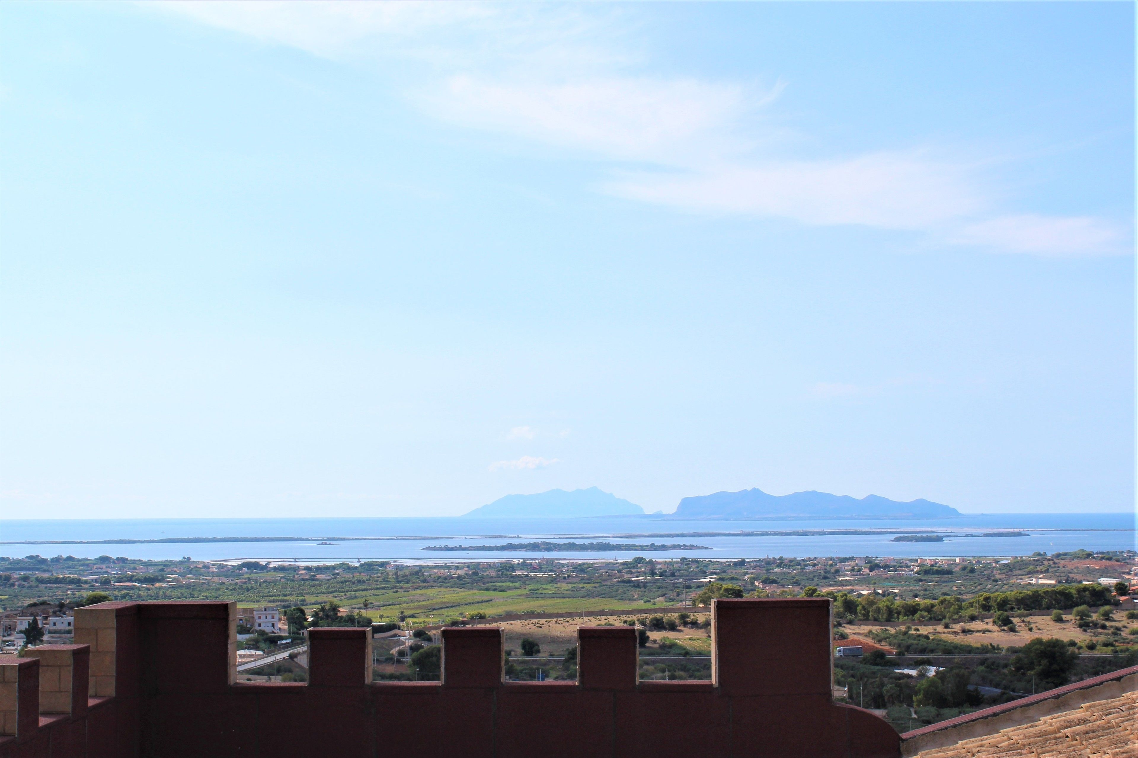 A coastal view from a high vantage point, overlooking a body of water, distant mountains, and a landscape with buildings and greenery.