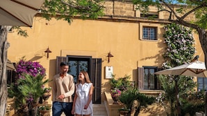 Una pareja está de pie frente a un edificio de fachada de piedra, con una puerta y un ventanal con contraventanas.