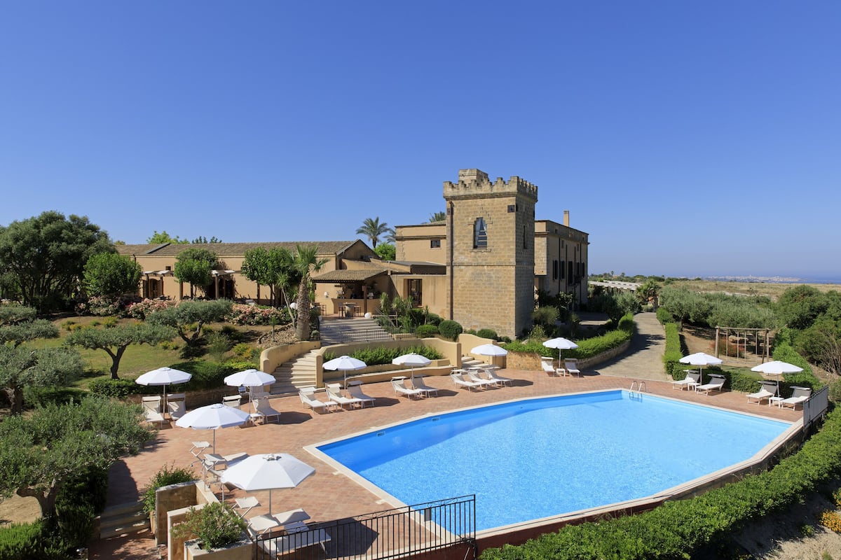 Una piscina con sillones de playa y sombrillas, rodeada de vegetación y un edificio con torre.