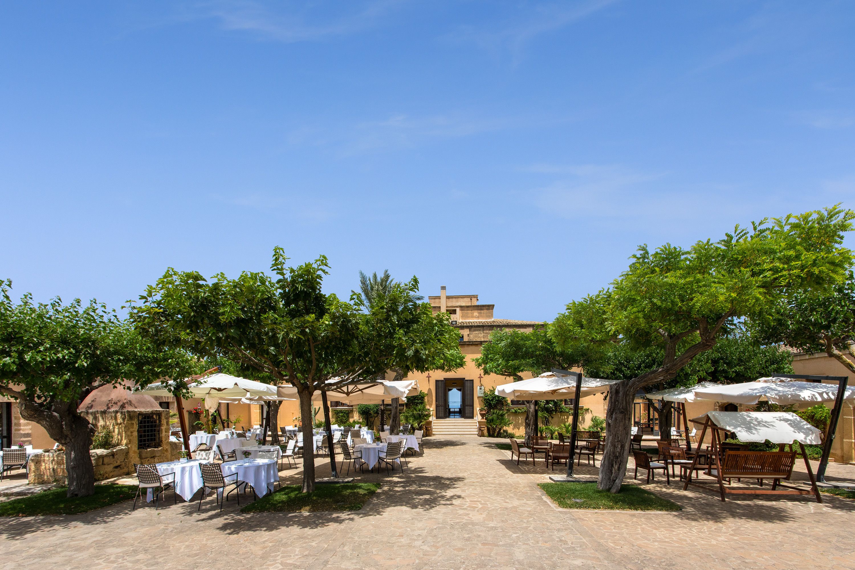 An outdoor dining area with tables and chairs under umbrellas, a stone pathway, and a building with a blue door in the background.