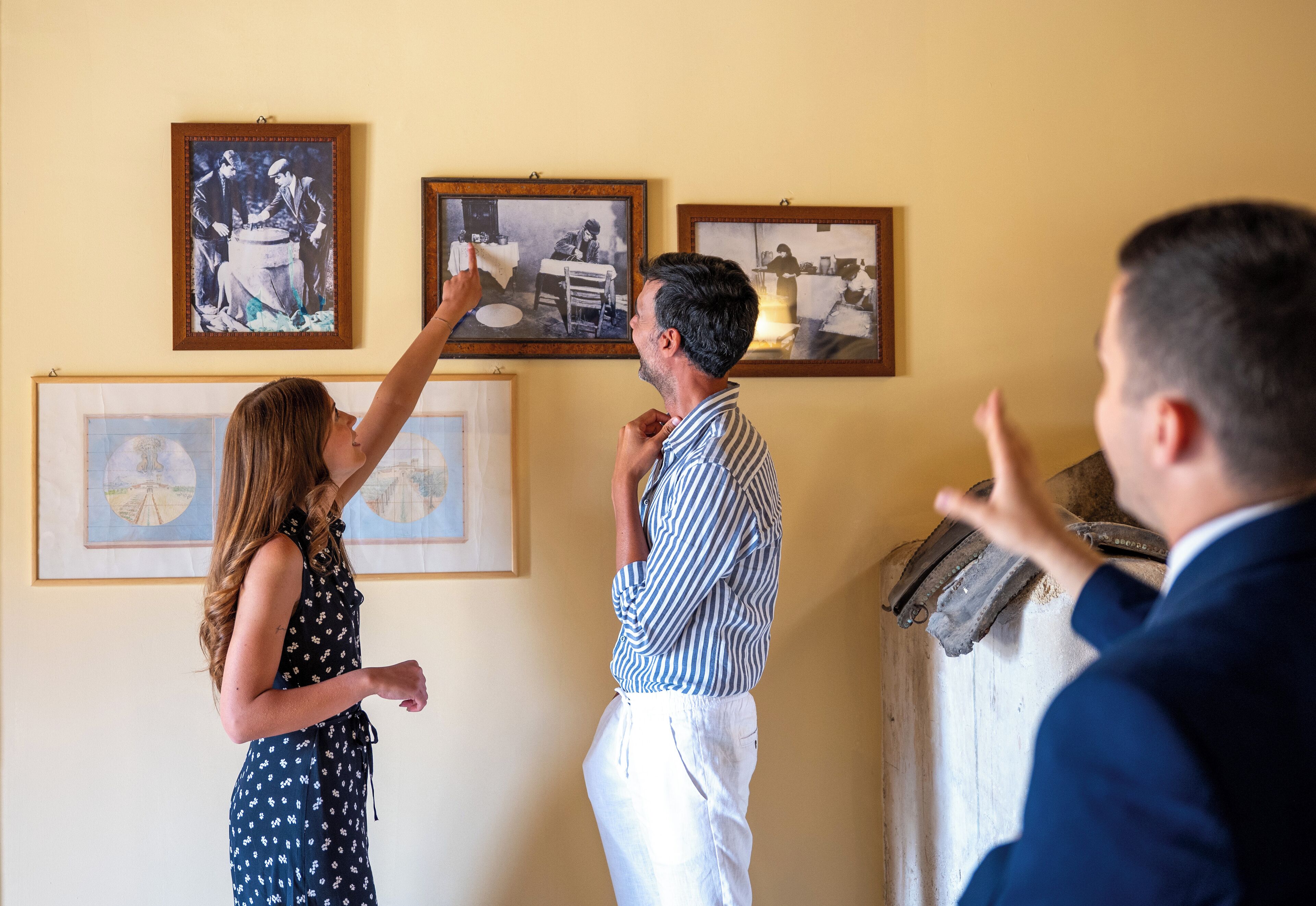 A room with framed pictures on the wall, a wooden cabinet, and a person pointing at the pictures.