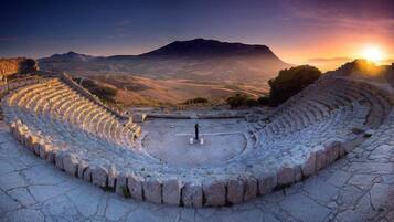 Un antico anfiteatro con posti a gradinate e un palcoscenico centrale, circondato da montagne e illuminato da un tramonto.