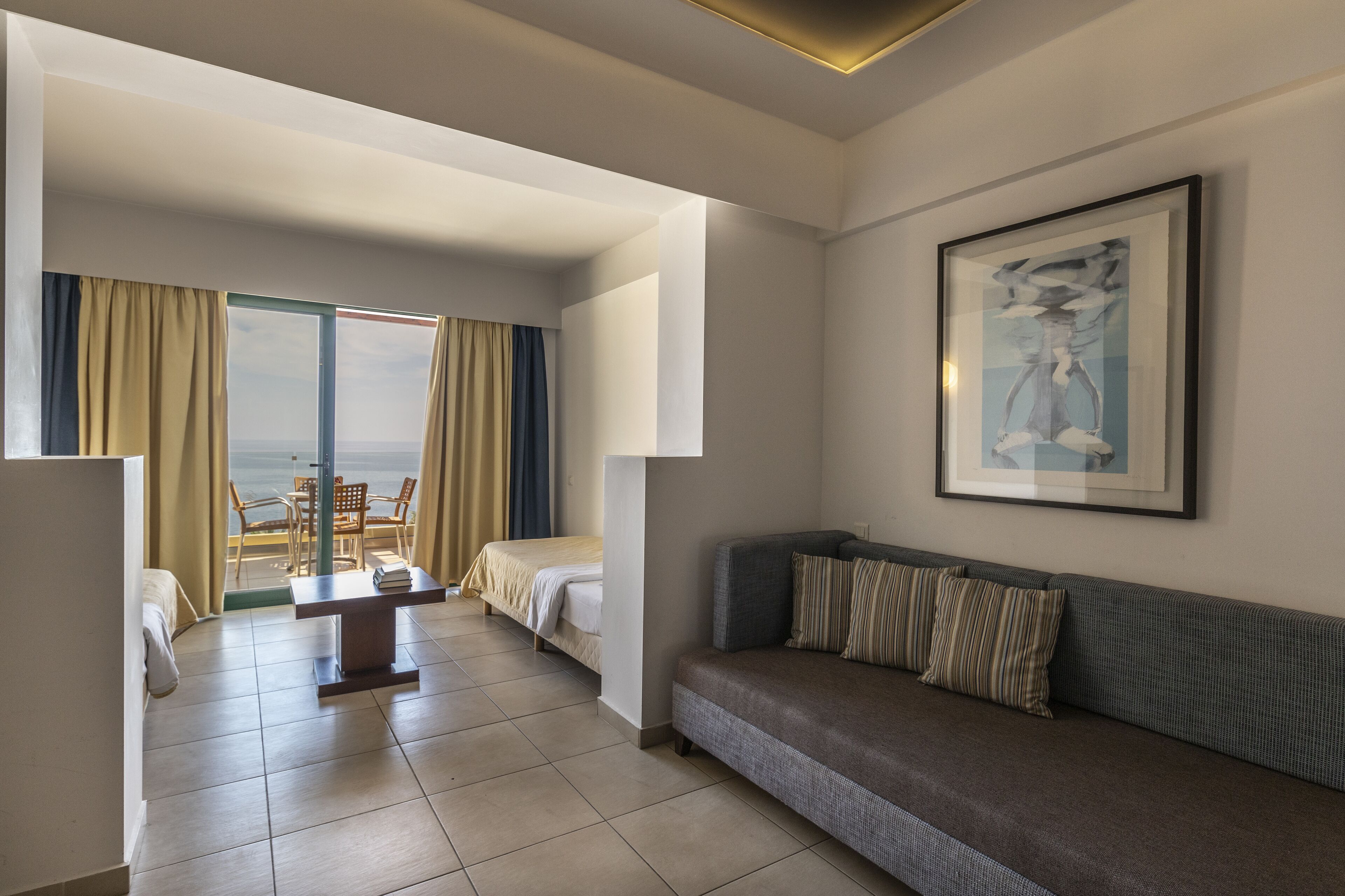 Quarto de hotel com cama, sofá, mesa de jantar e vista para o mar através de grandes janelas.