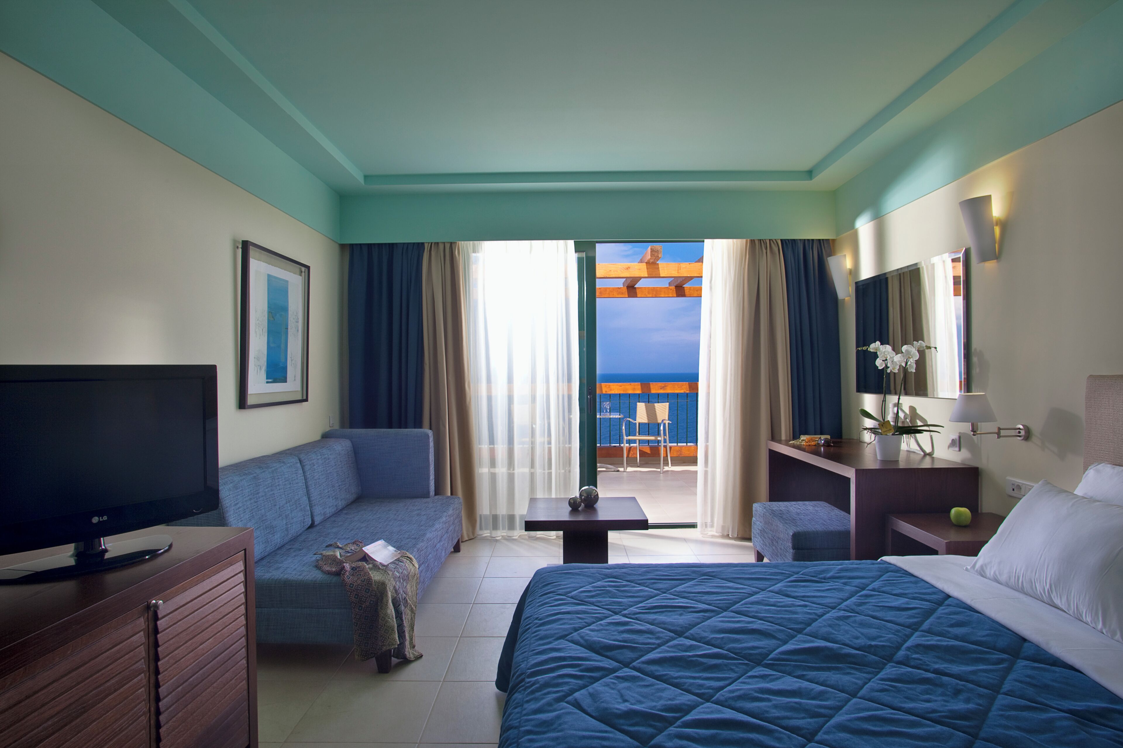 Ein Hotelzimmer mit Bett, Fernseher, Sofa, Schreibtisch und einem Balkon mit Meerblick.